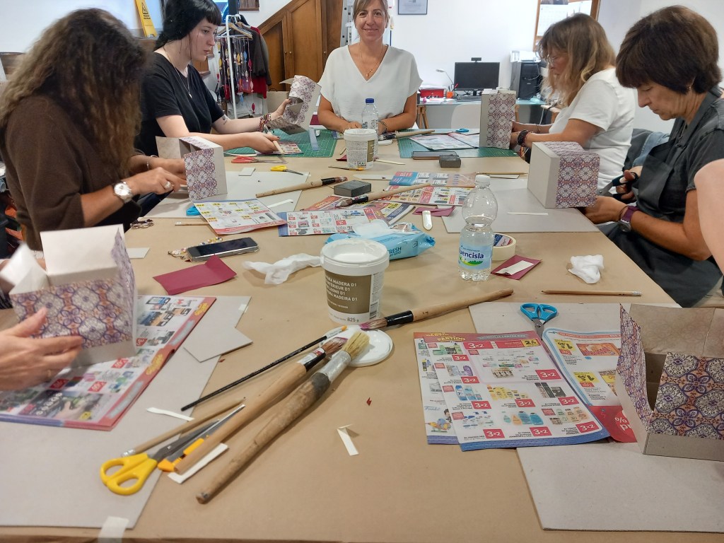Un grupo de mujeres participa en un taller de manualidades, trabajando en cajas decorativas en una mesa llena de materiales como tijeras, papel y herramientas.